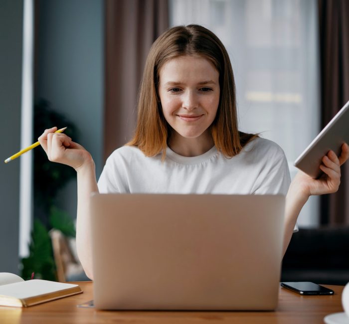 A smart student learns at home online via the internet. On a wooden table stands a laptop in the hands of a tablet. Distance learning at the Institute. Young red-haired woman freelancer.